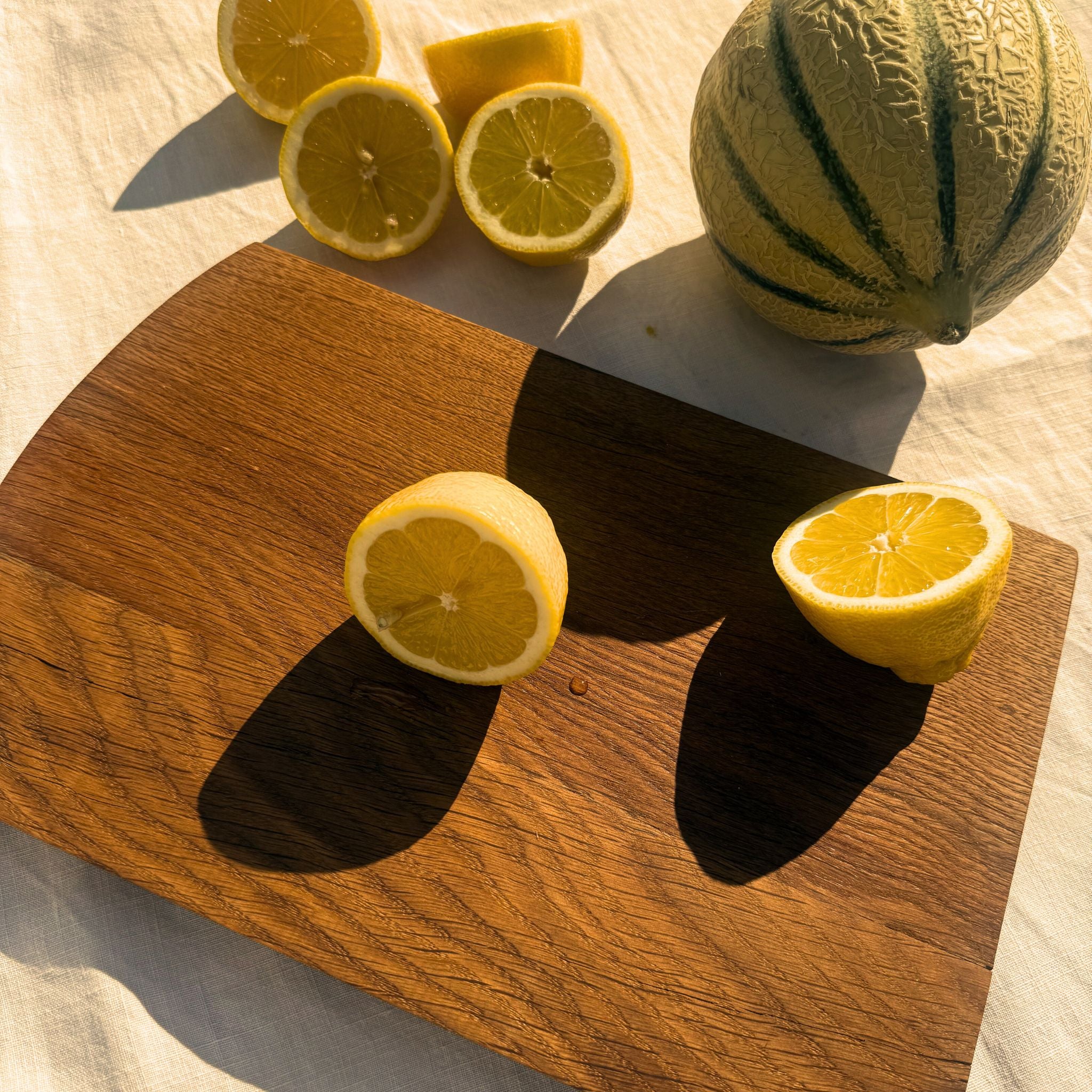 Planche à découper Aiona en chêne, avec citrons et melon, mise en lumière dorée du soleil.