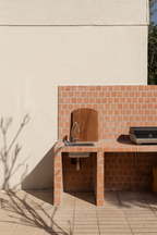 Planche à découper Aiona modèle CAVAILLON en chêne, accrochée sur un mur carrelé terracotta d’une cuisine extérieure avec barbecue.